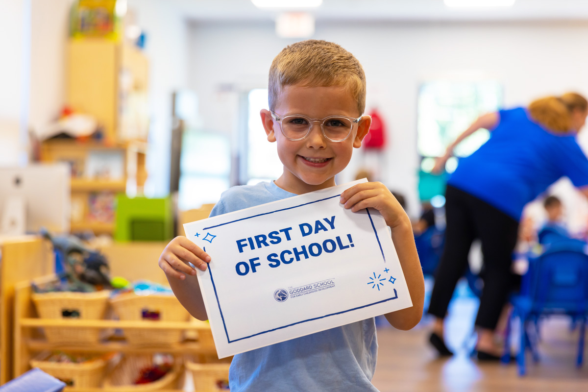 A young smiling boy in glasses holding a 'First Day of School!' sign in a daycare preschool classroom.