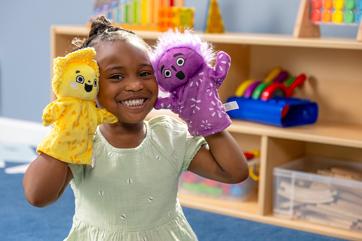 A joyful child in a light green dress holds a yellow and a purple puppet, smiling widely. Shelves with colorful toys are in the background, suggesting a playroom.
