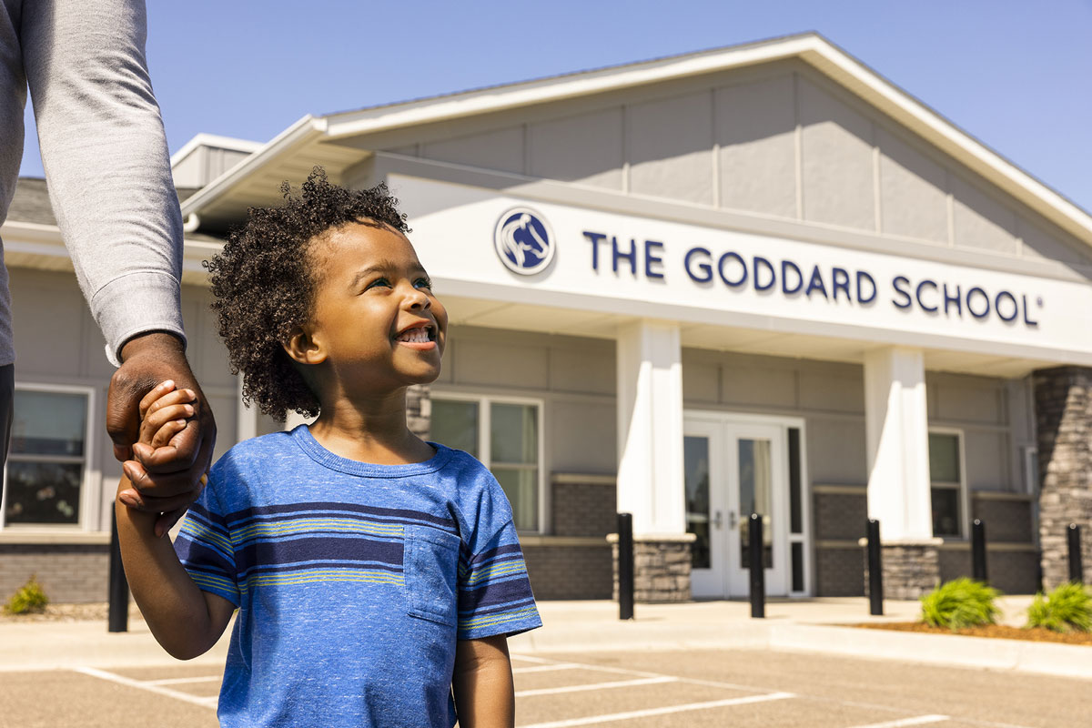 Young child holding his dad's hand smiling up at The Goddard School in the background