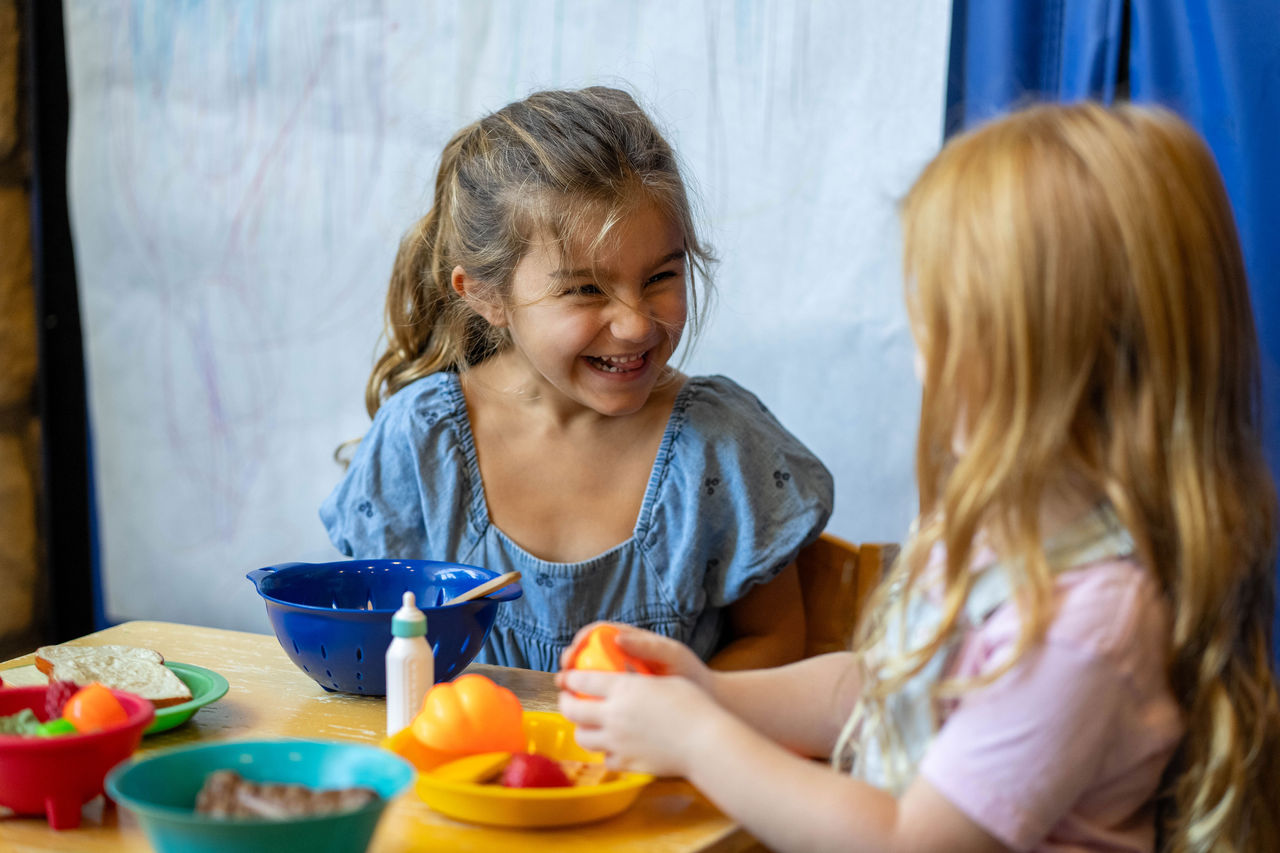 Two young girls laughing and chatting while playing with toy food at a table in a preschool classroom