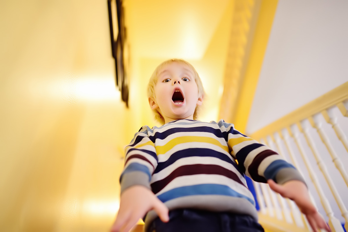 A young child wearing a striped sweater stands indoors, looking down with a wide open mouth and raised arms, appearing surprised or scared.